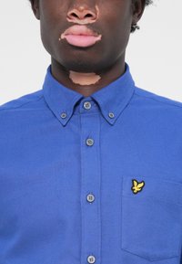 Blå skjorte med knapper og teksturert overflate, med brystlomme og brodert gult logo. Standard krage og grå knapper.