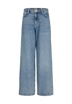Garçon debout, portant un t-shirt blanc à manches courtes, un jean bleu ample et des chaussures beiges, regardant vers sa droite sur un fond clair uni.