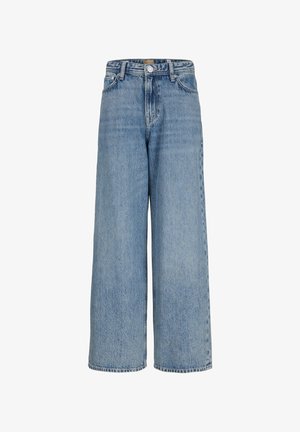 Garçon debout, portant un t-shirt blanc à manches courtes, un jean bleu ample et des chaussures beiges, regardant vers sa droite sur un fond clair uni.