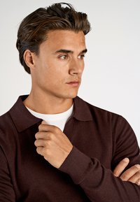 Ciemnoczerwony sweter z kołnierzykiem, długimi rękawami i ściągaczami. Nosi się go na białej koszulce, ukazując teksturowany materiał.