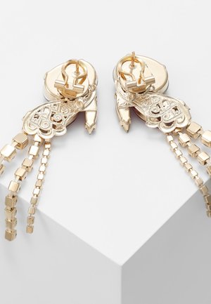 Goudkleurige clip-on oorbellen met filigraan details en drie hangende kettingen met kleine vierkante schakels, tentoongesteld op een wit voetstuk.