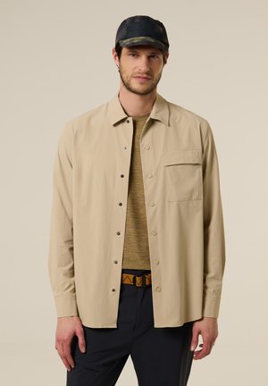 Homme portant une surchemise beige à boutons, un t-shirt vert olive, un pantalon noir, une ceinture marron à motif et une casquette noire, sur un fond uni.