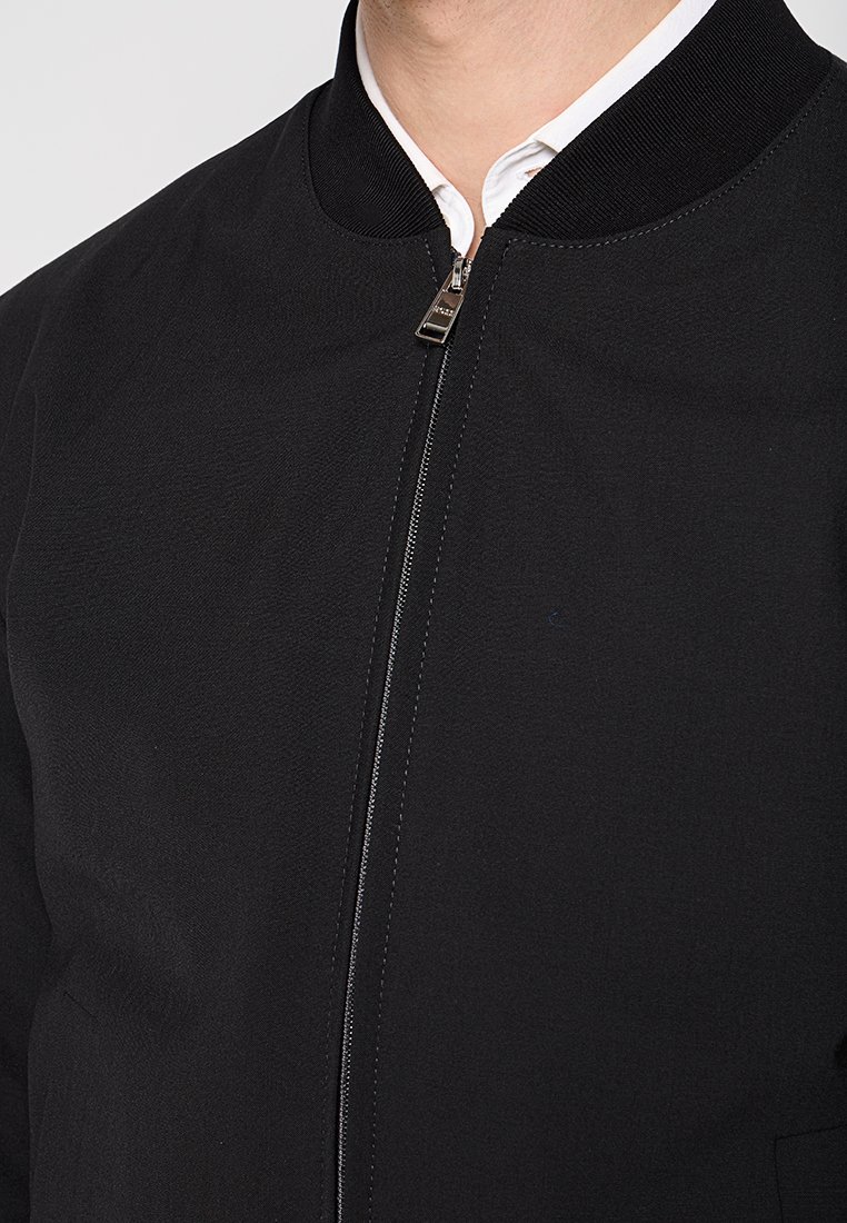 Schwarze Bomberjacke aus glattem Material, mit geripptem Kragen und einem Reißverschluss vorne. Besitzt minimale Details und eine taillierte Passform.