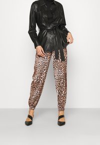 Chemise enveloppante en cuir noir à manches longues, associée à un jogger imprimé léopard aux nuances de brun et beige. Des ballerines noires pointues complètent la tenue.