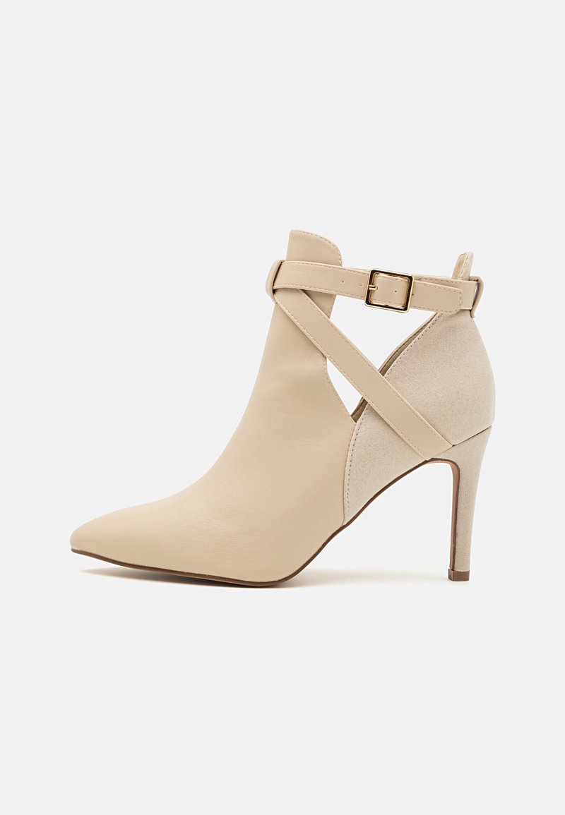 Bottine beige à talon haut et bout pointu avec arrière en daim, brides croisées à la cheville et boucle dorée sur un fond blanc uni.