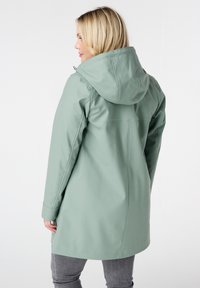 Lichtgroene regenjas met capuchon, met een strak ontwerp, gladde textuur en gestikte details. Draagt boven grijze broek.