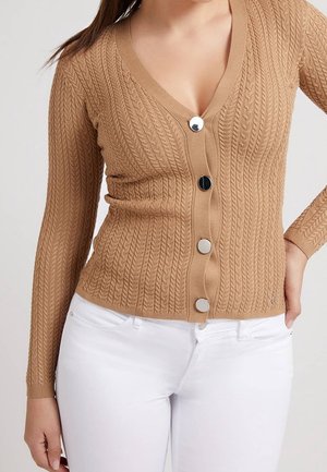 Bronzefarbene Strickjacke mit V-Ausschnitt, langen Ärmeln und drei silbernen Knöpfen auf der Vorderseite. Kombiniert mit weißen Hosen.