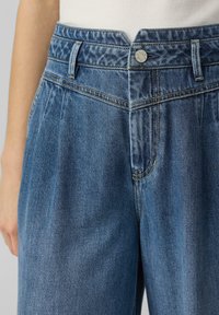 Pantalones de mezclilla de cintura alta, color azul claro, con pliegues frontales y un detalle de botón plateado. La textura de la tela es suave con un brillo sutil.