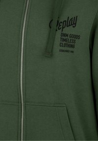 Felpa con zip di colore verde scuro in tessuto testurizzato, con un logo e testo stampati in nero e un cordino sul cappuccio.