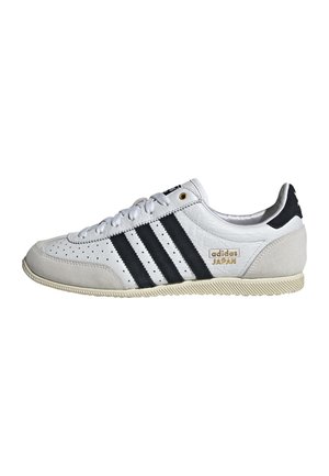 Witte Adidas Japan sneaker met zwarte strepen, beige accenten, witte veters en een gestructureerde zool, van opzij getoond op een witte achtergrond.