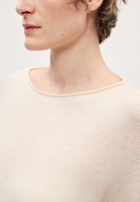 Beżowy sweter z dzianiny o szerokim, zaokrąglonym dekolcie i subtelnej fakturze. Materiał wydaje się miękki i lekki.