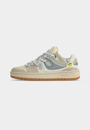 Zapatilla beige y gris con cordones gruesos blancos y grises, suela de goma y un detalle de cara sonriente amarilla en el lateral cerca del talón.