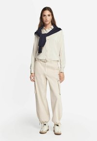 Camicia bianca a righe con bottoni, pantaloni cargo beige con polsini regolabili, maglione blu navy legato attorno al collo e scarpe da ginnastica bianche con dettagli beige.