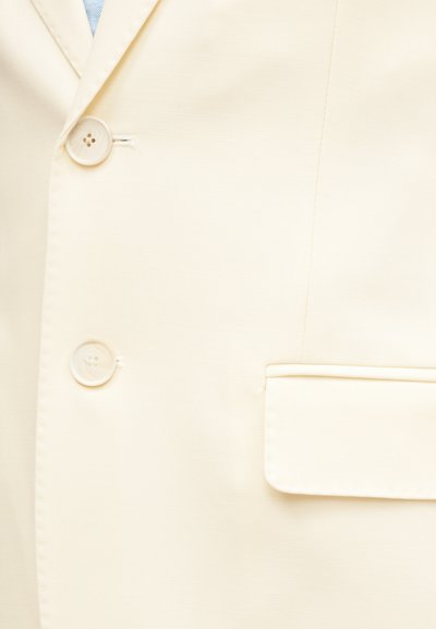 Κοντινό πλάνο εμπρός μέρους σακακιού σε κρεμ απόχρωση με δύο κρεμ κουμπιά, τσέπη με καπάκι στην δεξιά πλευρά και μια διακριτική υποψία ανοιχτού μπλε πουκαμίσου από κάτω.