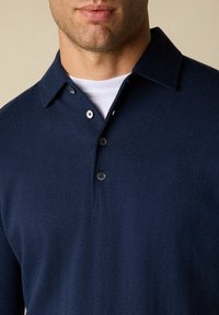 Námořnická modrá polo košile vyrobená z měkké, strukturované tkaniny. Obsahuje límec s třemi knoflíky a bílou vnitřní vrstvu viditelnou na límci.