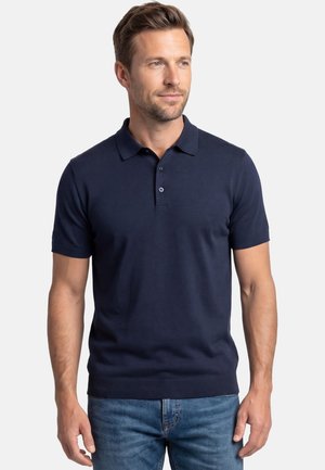 Mies, jolla on tummansininen lyhythihainen poolopaita ja siniset farkut, seisoo yksinkertaista vaaleaa taustaa vasten ja katsoo hieman sivulle.