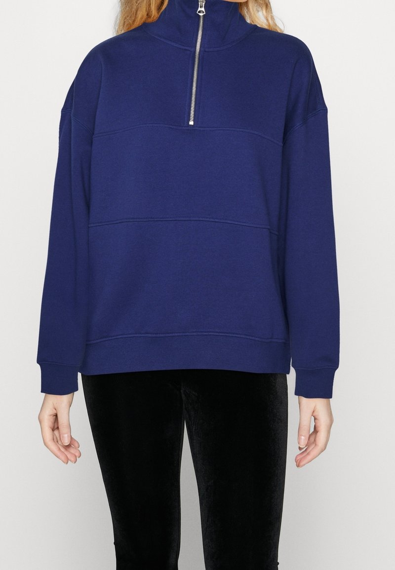 Personne portant un pull bleu marine avec un col montant et une demi-fermeture éclair argentée, associée à un pantalon en velours noir, sur un fond uni.