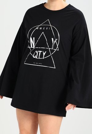 Zwart oversized shirt met lange mouwen met zilveren geometrische vormen en tekst "NY CITY" gedragen door een persoon met blote benen zichtbaar.