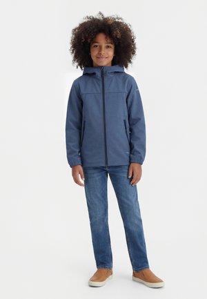 Copil cu păr creț stă zâmbind, purtând o jachetă albastră cu glugă, blugi albaștri și pantofi slip-on bej, pe un fundal alb.