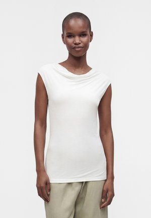 Vrouw met kort haar die een wit mouwloos topje en lichtbeige broek draagt, staand tegen een effen lichte achtergrond.