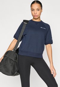 Σκούρο μπλε κοντομάνικο φούτερ με στρογγυλή λαιμόκοψη και υφασμάτινο τελείωμα, συνδυασμένο με μαύρο κολάν και γυμναστηριακή τσάντα με διακριτικά από μαύρο δίχτυ.