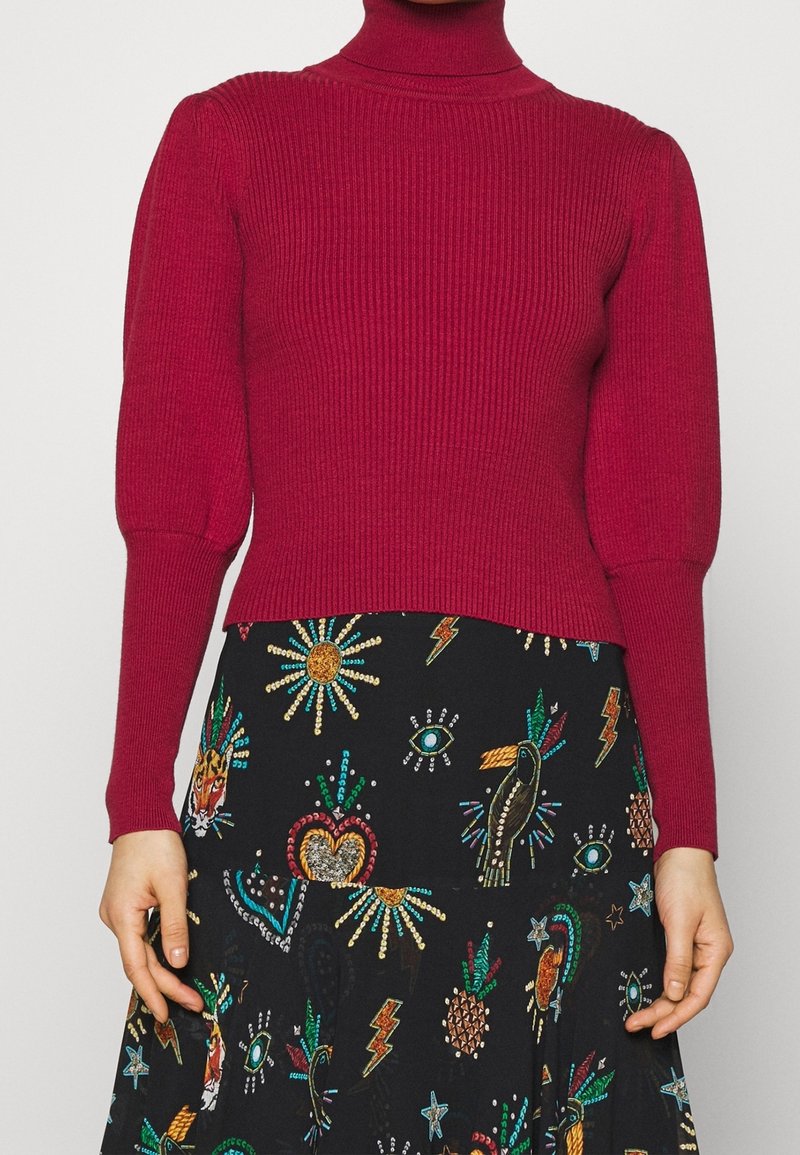 Personne portant un pull à col roulé rouge à manches longues, en maille côtelée, et une jupe noire ornée de motifs brodés colorés représentant des yeux, des oiseaux, des cœurs et des étoiles.