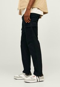 Pantaloni cargo in denim blu scuro con vestibilità rilassata, dotati di molteplici tasche e una texture liscia. Abbinati a sneakers grigie e bianche.