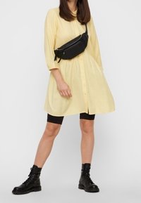 Gelbes Hemdkleid mit Knöpfen, knielang mit langen Ärmeln. Schwarze Bauchtasche am Taillengürtel getragen und schwarze Kampfstiefel. Schwarze Leggings sichtbar.