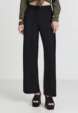 Schwarze weite Hosen aus einem glatten, fließenden Stoff. Mit Falten an der Vorderseite und kombiniert mit Plateausandalen.