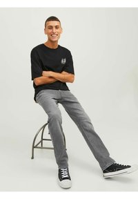 Νεαρός άνδρας που χαμογελά, φορώντας μαύρη μπλούζα με έμβλημα, γκρι τζιν και μαύρα αθλητικά παπούτσια, καθισμένος σε μεταλλική καρέκλα με τα χέρια σταυρωμένα.