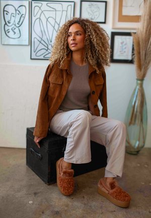 Veste en velours côtelé marron, haut beige, pantalon clair et chaussons marron duveteux posés sur une boîte noire. En arrière-plan, une œuvre d'art abstraite.