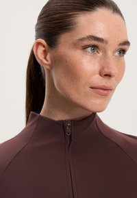 Burgundi színű, testhez simuló dzseki magas gallérral és cipzáras zárással. Simább anyag, amely karcsú dizájnnal emeli ki a nyakat és a vállakat.