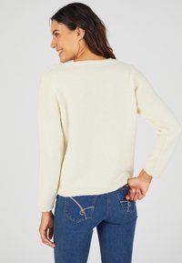 Pull en tricot couleur crème avec des manches longues, ourlet côtelé et texture douce, associé à un jean bleu foncé avec des coutures décoratives sur les poches.