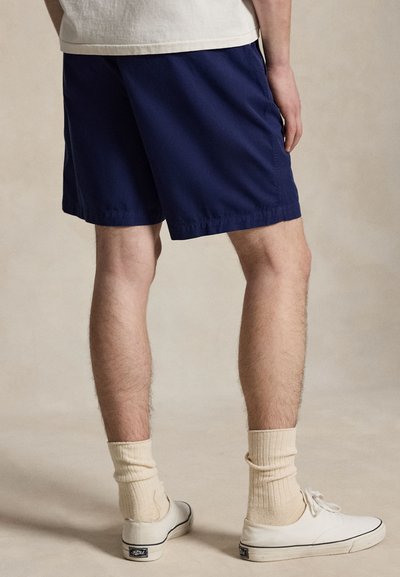 Muž v tmavomodrých kraťasoch, krémových ponožkách a bielych ležérnych topánkach stojaci s mierne rozšírenými nohami pred jednofarebným béžovým pozadím.