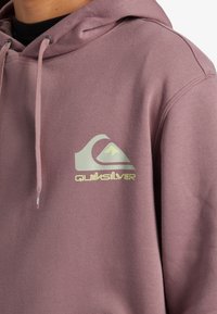 Hooded sweatshirt in mauve, featuring a thick texture. Front shows a white and green Quiksilver logo accent. Drawstrings with silver tips.