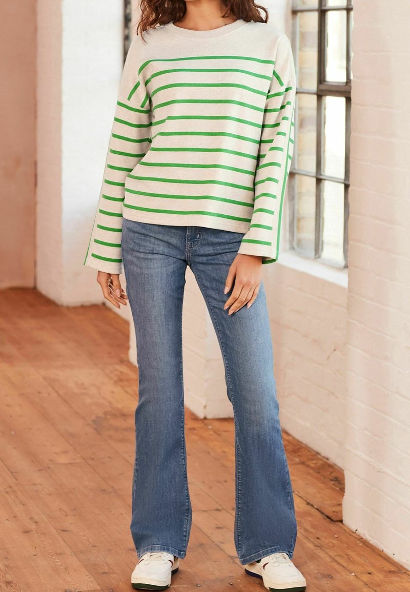Personne portant un pull crème avec des rayures horizontales vertes, un jean flare bleu et des baskets blanches, debout près d'une fenêtre sur un sol en bois.