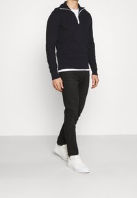 Granatowy sweter na zamek błyskawiczny z białym akcentem zamka, w połączeniu z czarnymi dżinsami i białymi sneakersami. Sweter ma prążkowane mankiety i dół.