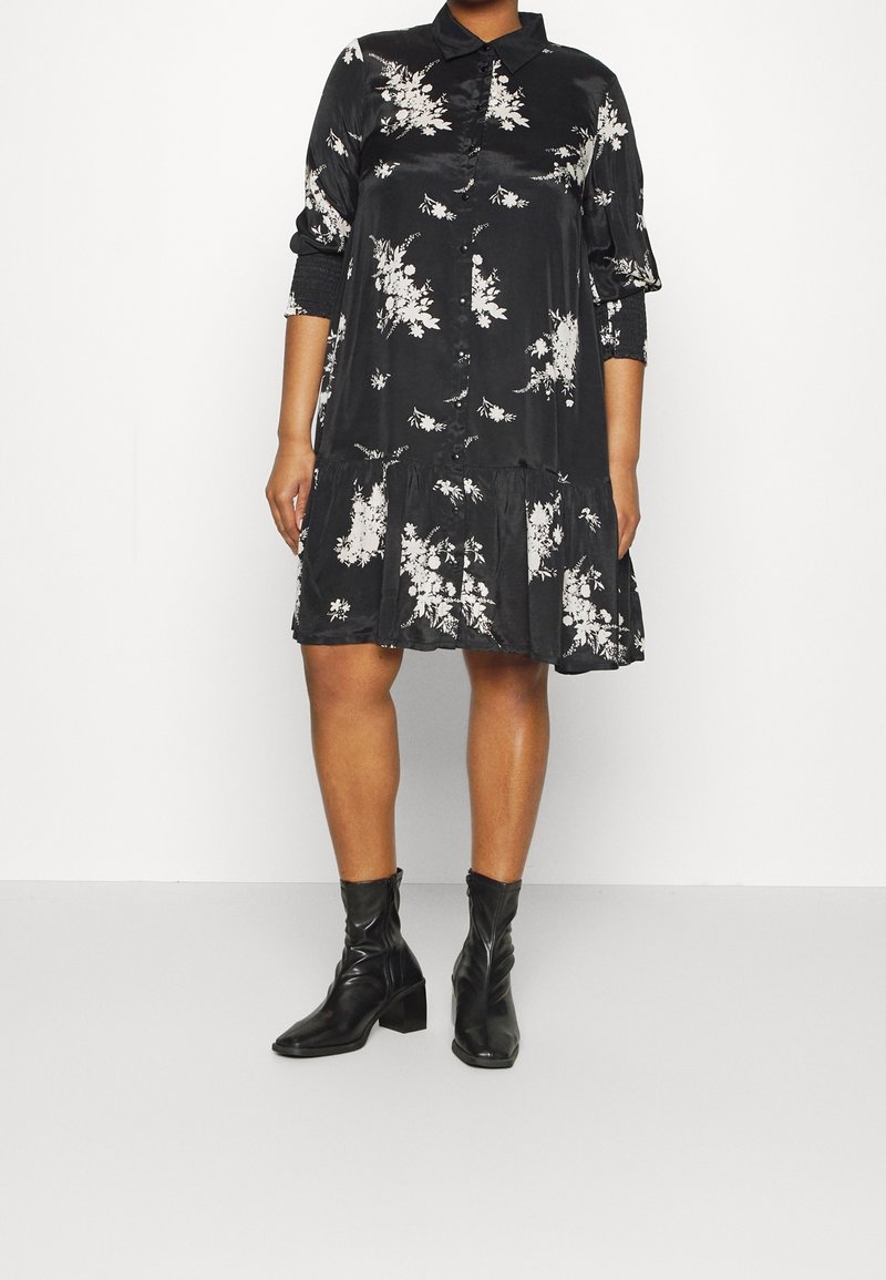 Femme portant une robe chemise noire à manches longues avec un imprimé floral, associée à des bottes noires mi-mollet à talons, debout devant un fond uni.