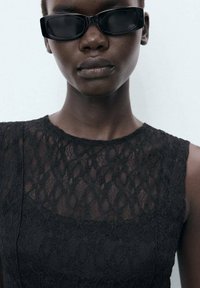 Top sans manches en dentelle noire avec des détails de motifs complexes ; modèle portant des lunettes de soleil sombres et angulaires. Fond uni.