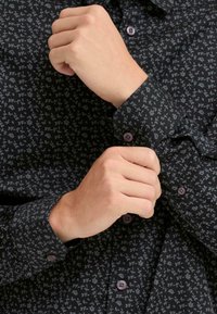 Camicia nera con bottoni a pressione, caratterizzata da un piccolo motivo floreale grigio. Presenta un colletto e polsini tradizionali, con bottoni coordinati. Tessuto strutturato.