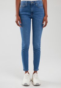 Sötét bőrű álló személy által viselt, közép kék színű, vékony fazonú farmerek, fehér tömzsi sportcipőkkel párosítva, sima világos háttéren.