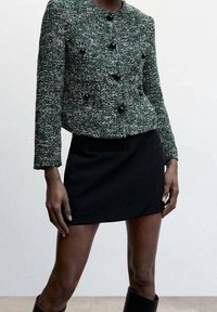 Veste en tweed vert et blanc avec détails de boutons noirs, manches longues, associée à une mini-jupe noire, texture lisse, forme ajustée.