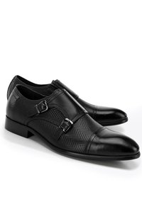Zapatos de vestir negros con parte superior texturizada, que presentan dos hebillas laterales, una puntera de cuero lisa y un tacón bajo apilado.