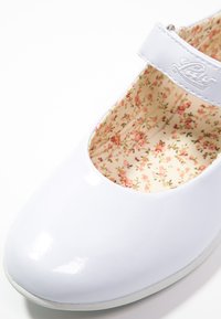 Witte lakleren schoen met een ronde neushak en verstelbare band, voorzien van een voering met een bloemmotief in zachte pastelkleuren.