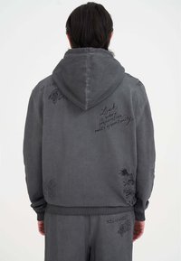 Sudadera gris con capucha ajustable, que presenta patrones bordados en negro de dragones y texto. Fabricada con un tejido texturizado y puños acanalados.