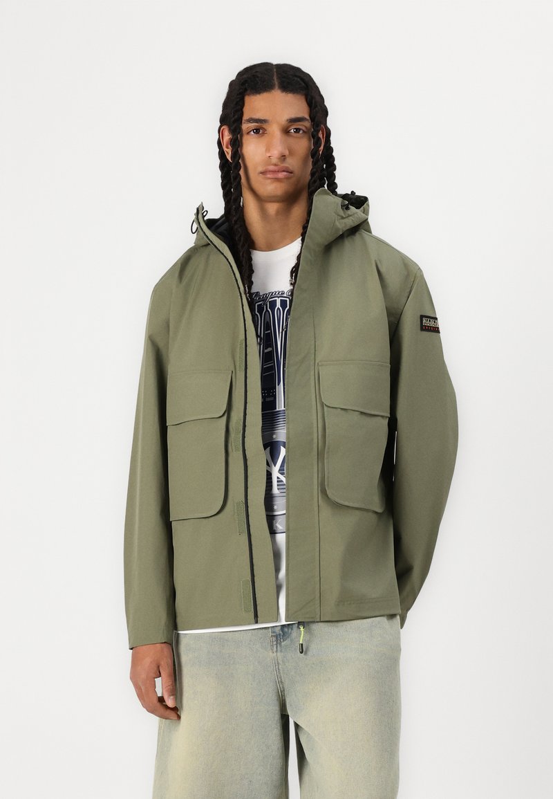 Chaqueta de color verde oliva claro, fabricada en un material impermeable. Presenta una capucha, dos bolsillos frontales y un cierre de cremallera con un zipper en contraste.
