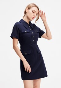 Robe bleu marine à manches courtes au design ajusté avec un bouton sur le devant, deux grandes poches poitrine et un col pointu. Texture de tissu lisse.