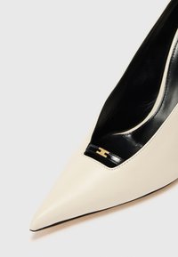 Béžový kožený stiletto podpätok s špičatou špičkou, výrezom, čiernym interiérom a malým zlatým kovovým detailom. Hladká textúra.