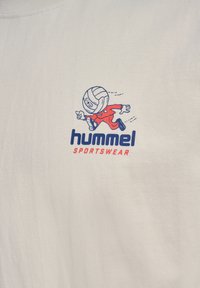 Biely bavlnený tričko s potlačou rozprávkového charakteru s hlavou volejbalovej lopty v červenom oblečení. Pod tým je potlačené "hummel SPORTSWEAR" v modrej a červenej farbe.