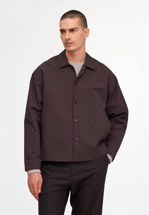Man draagt een donkerbruine gebuttonde jas over een grijze trui en zwart pantalon met fijne strepen, staand tegen een eenvoudige witte achtergrond.